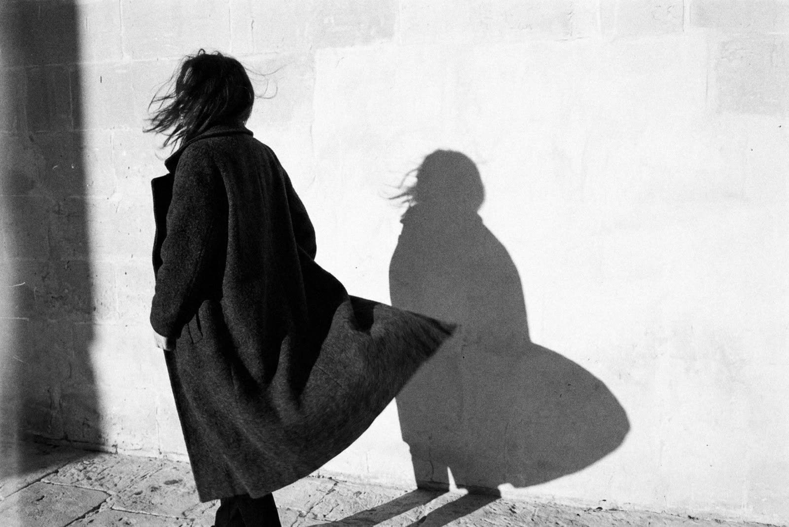 A woman in a coat in the wind, her shadow on a white wall
